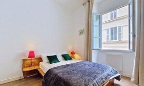 A bedroom with a bed, two bedside lamps, a window with curtains, and a radiator. - N&J - BLEU PANORAMA - Sea View (Niza)