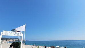 Eine Strandansicht mit einem Schild, auf dem 'LE TEMPS D'UN ÉTE' steht, und einem strahlend blauen Himmel.