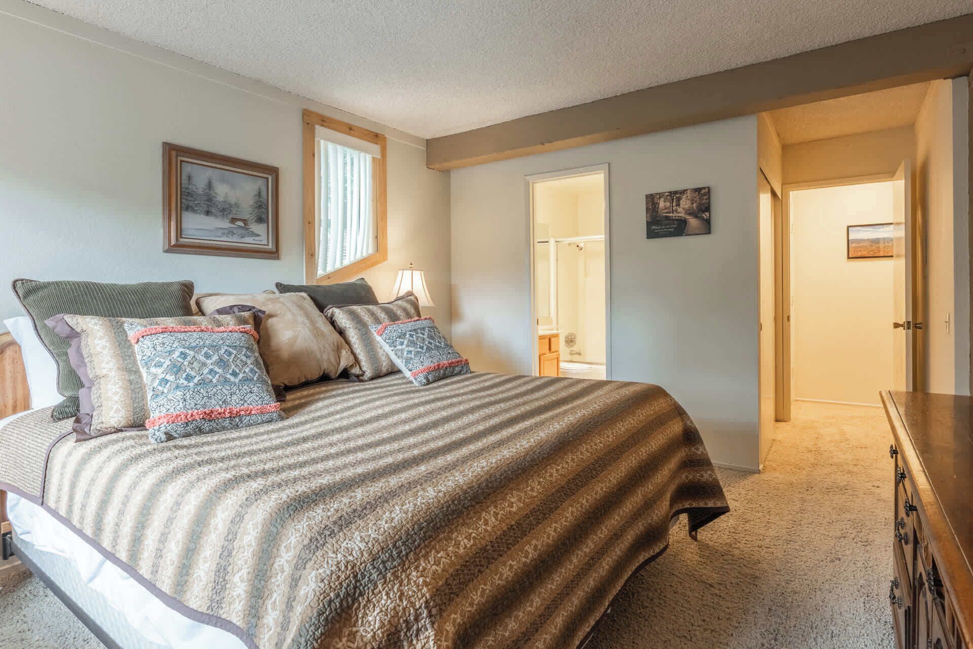 A bedroom with a large bed, a wooden headboard, a nightstand, a dresser, and a door leading to a bathroom.