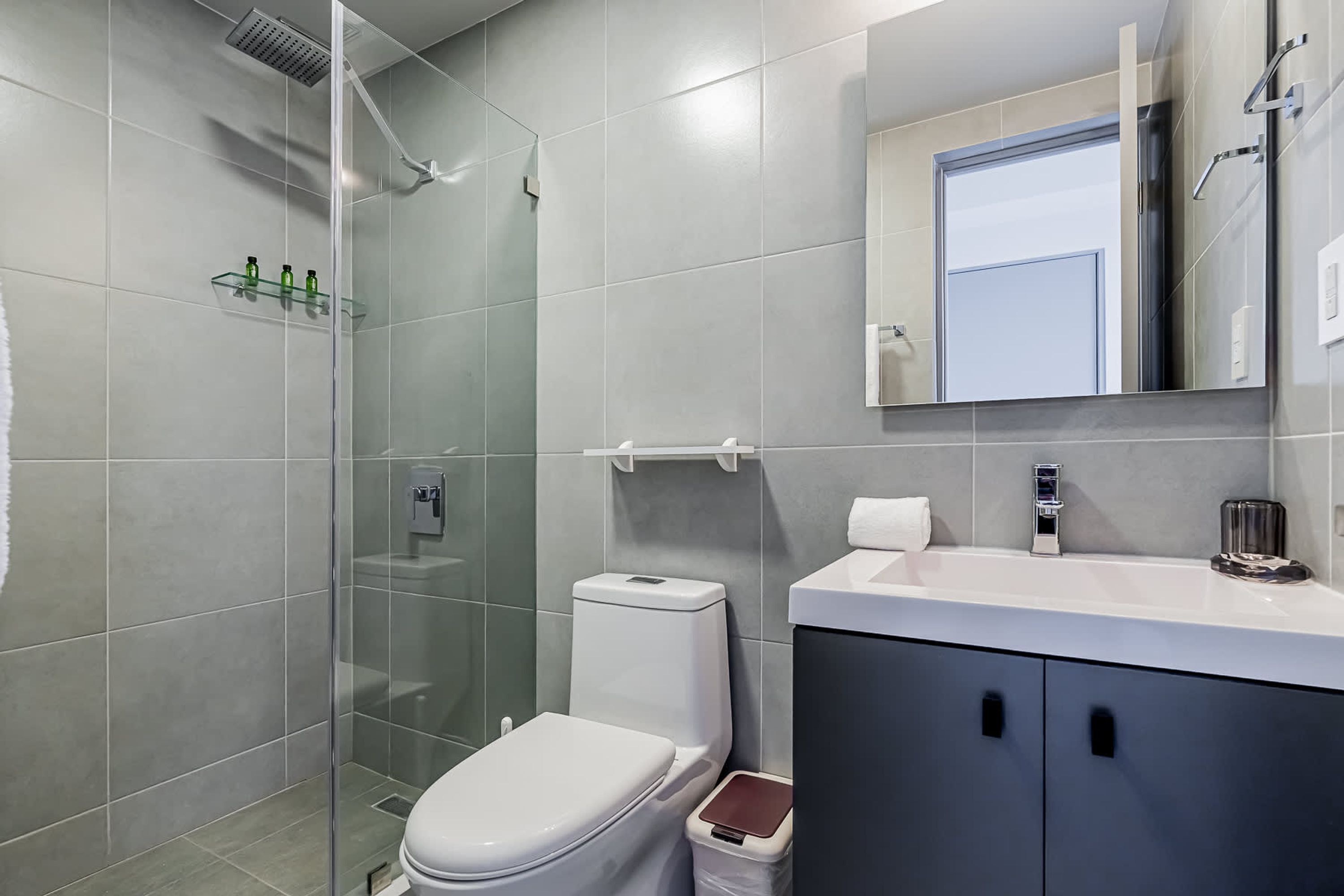 Un baño moderno con cabina de ducha de vidrio, inodoro blanco, tocador con espejo y un cubo de basura.
