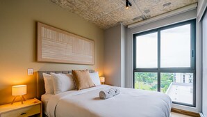 Een netjes opgemaakt bed met een opgerolde handdoek, een bedlamp en een groot raam met uitzicht op gebouwen en groen.