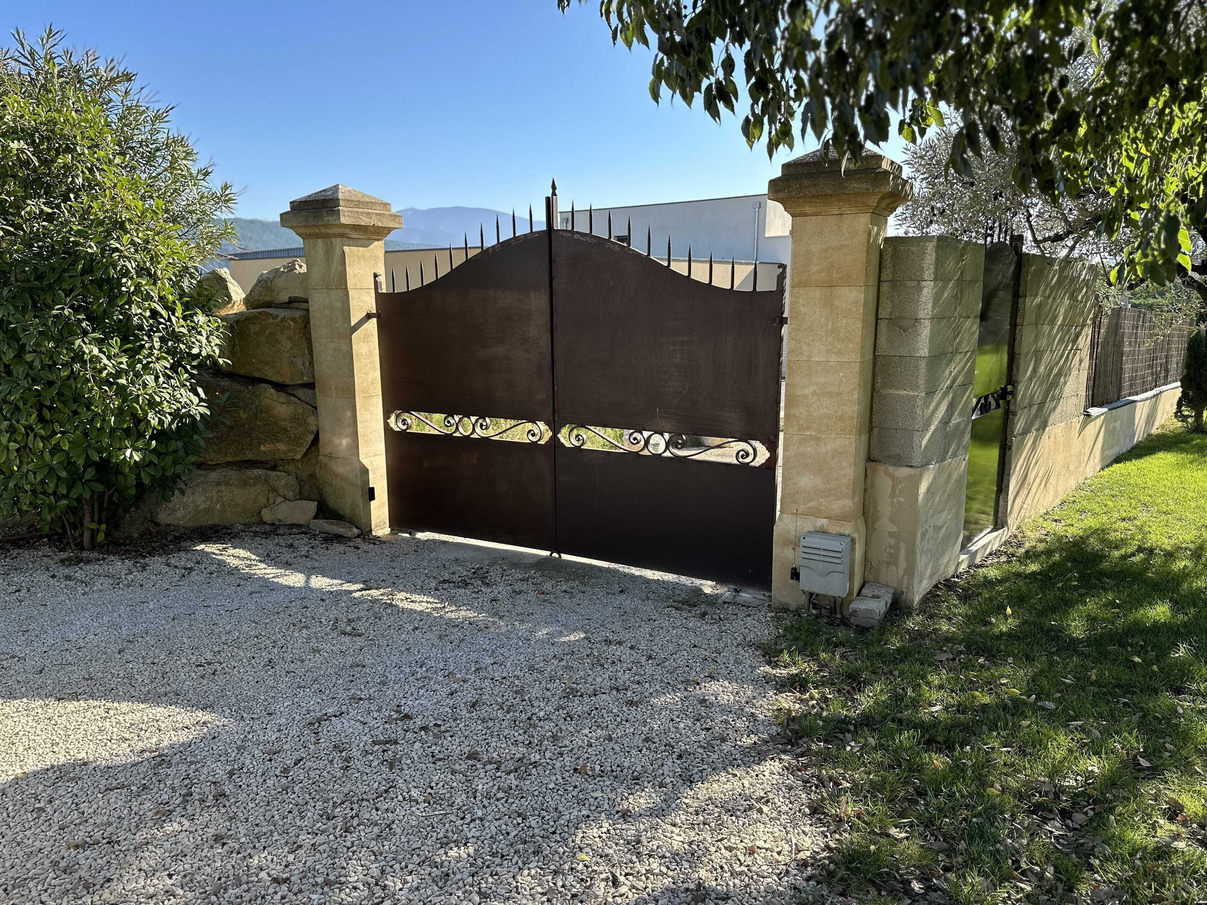 Une entrée fermée par un portail en métal décoratif, flanquée de piliers en pierre et précédée d’une allée gravillonnée.