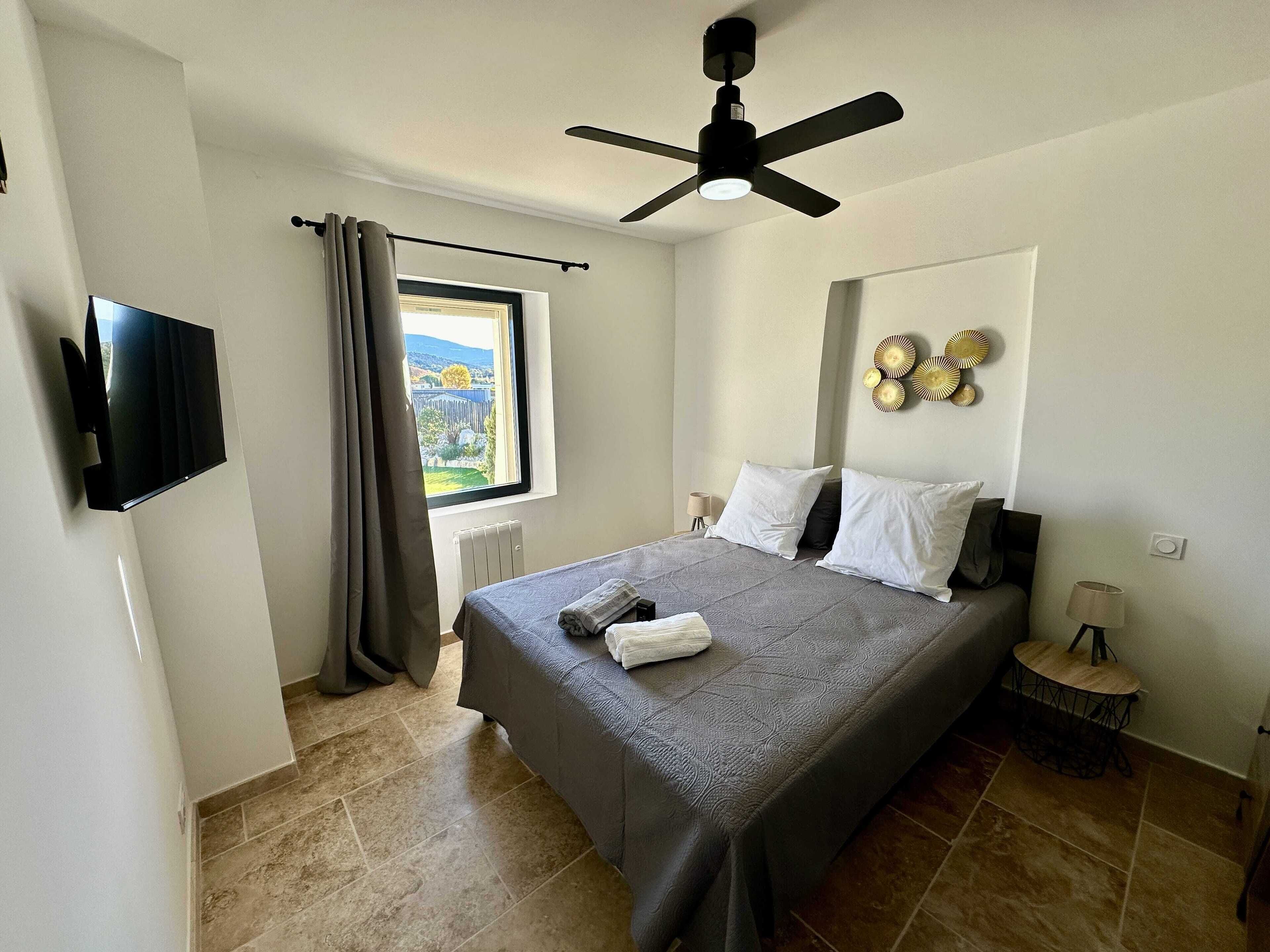 Una camera da letto con un letto, una ventola a soffitto, una TV fissata al muro, una finestra con tende, un termosifone e un comodino con una lampada.
