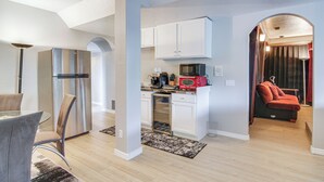 Eine moderne Küche mit weißen Schränken, einem Edelstahlkühlschrank und einer roten Mikrowelle. Es gibt einen Essbereich mit einem Glastisch und Stühlen, und im Wohnzimmer steht ein rotes Sessel.