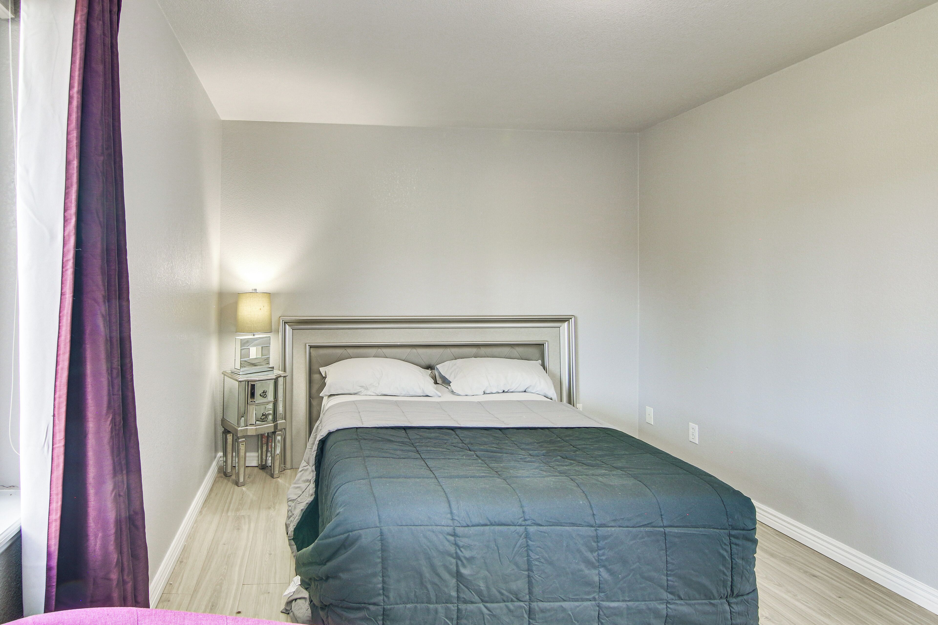 A neatly made bed with a dark blue quilt, a bedside table with a lamp, and a window with purple curtains.