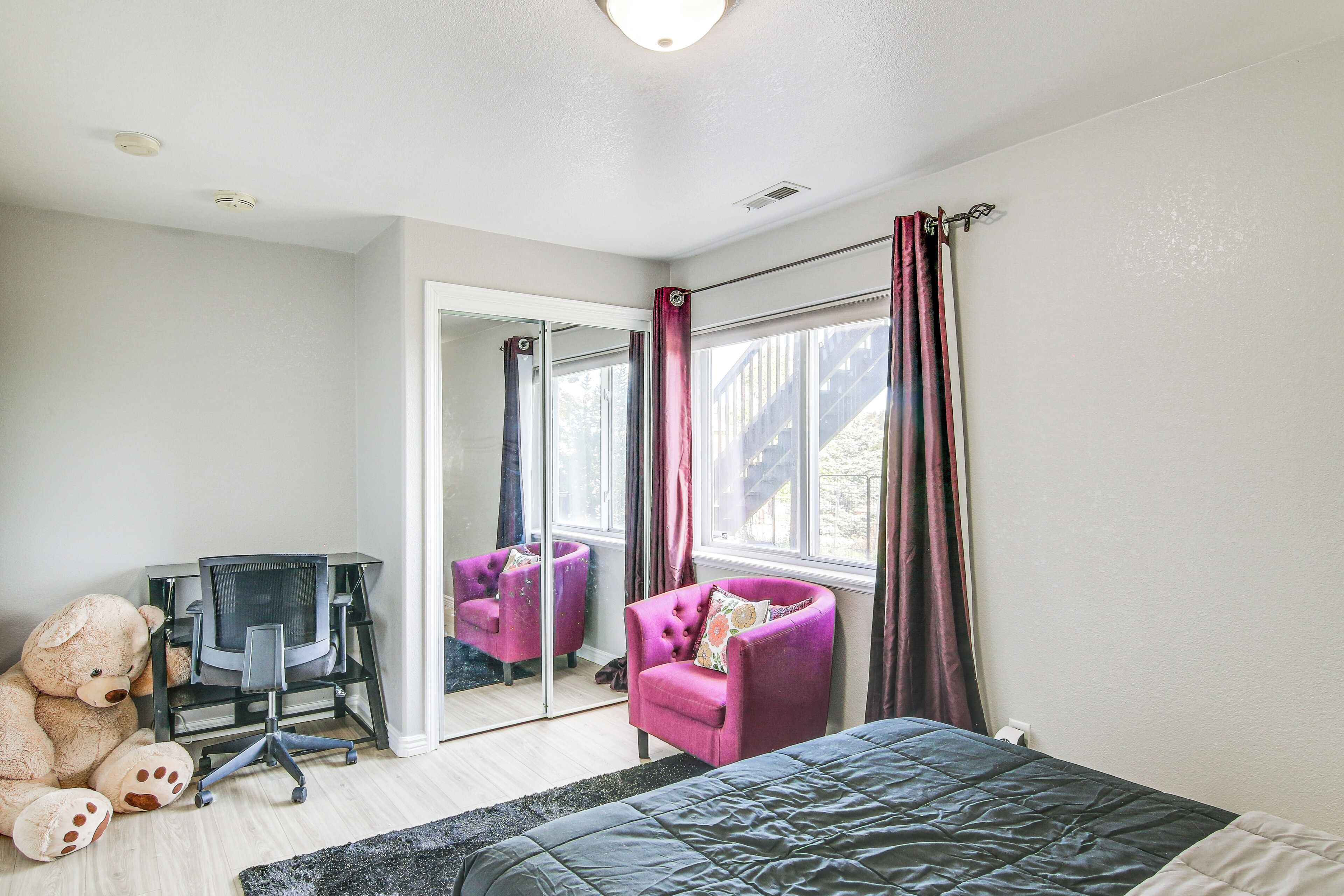 A bedroom with a bed, a desk with a chair, a large teddy bear, and a window with curtains.