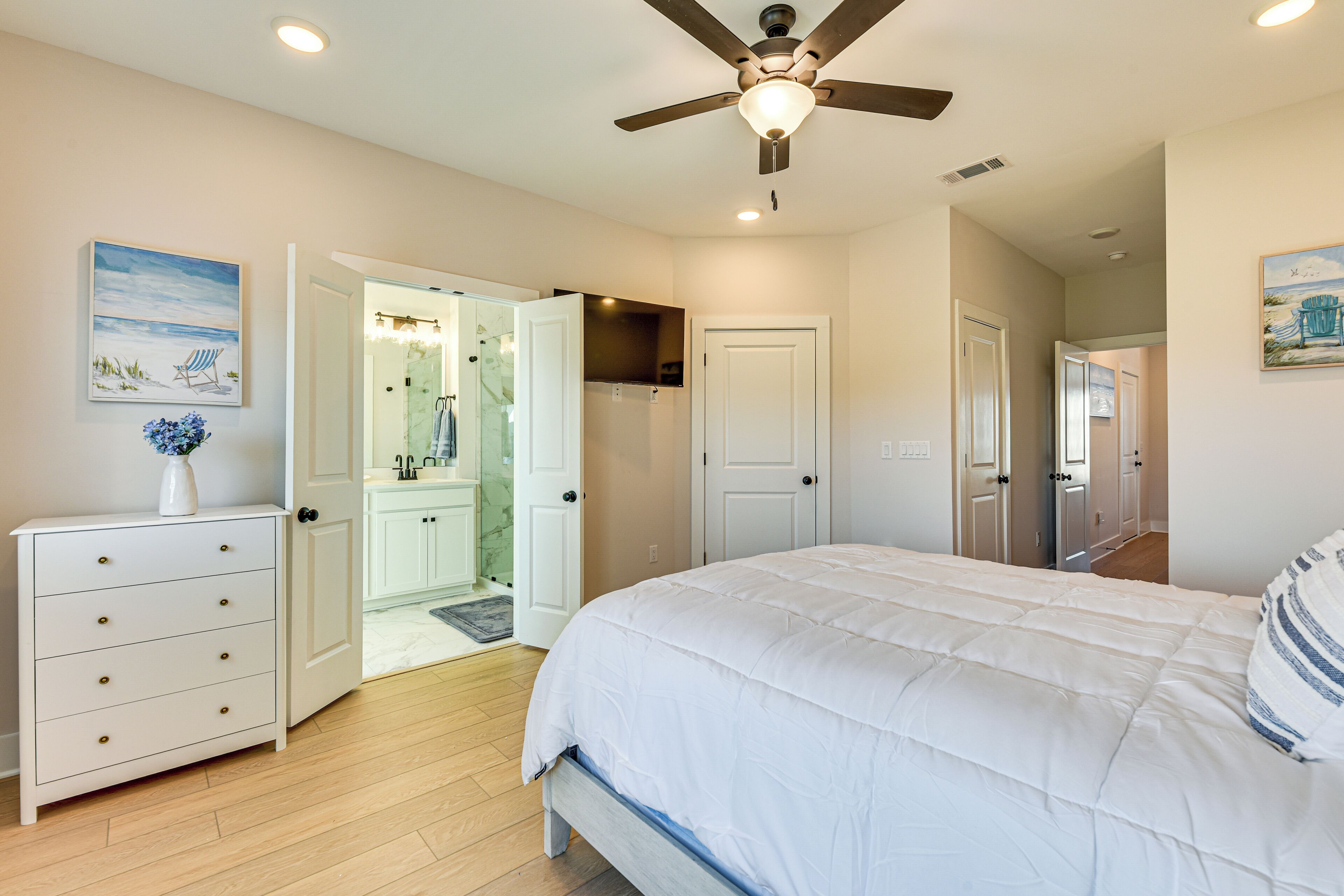 Une chambre à coucher avec un lit, une commode, un ventilateur de plafond et une salle de bain visible à travers une porte ouverte.