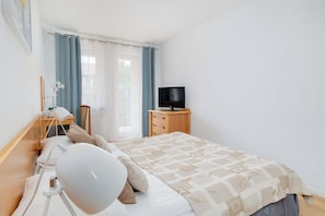 A bedroom with a bed, a bedside table with a lamp, a TV on a wooden stand, and a window with curtains.