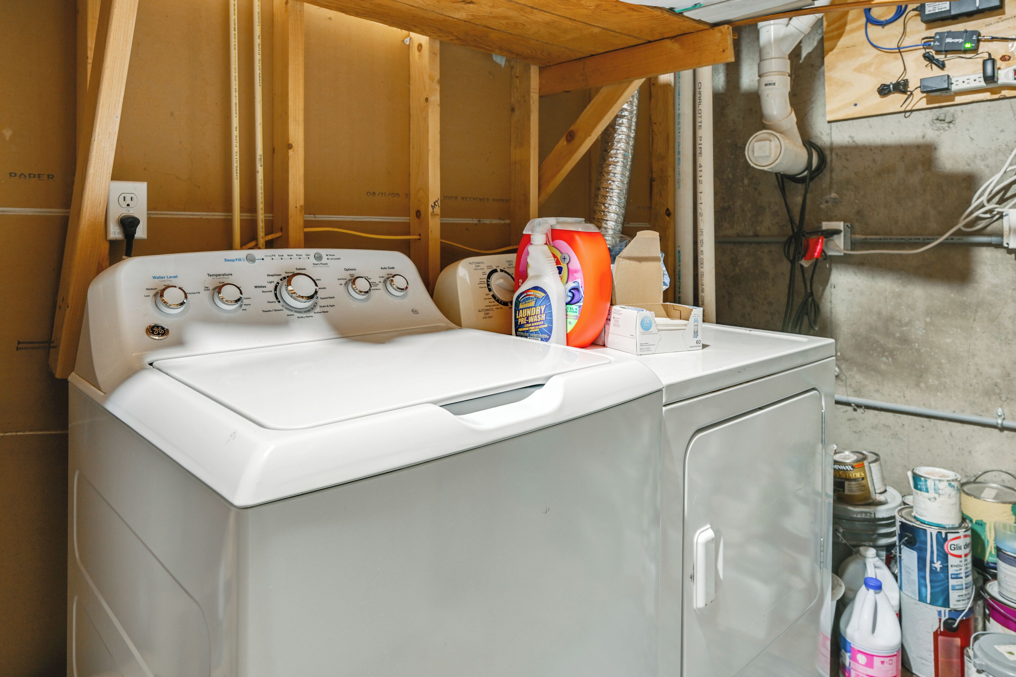 A white front-loading washing machine with control knobs, a bottle of laundry detergent, and various cleaning supplies on a wooden floor.