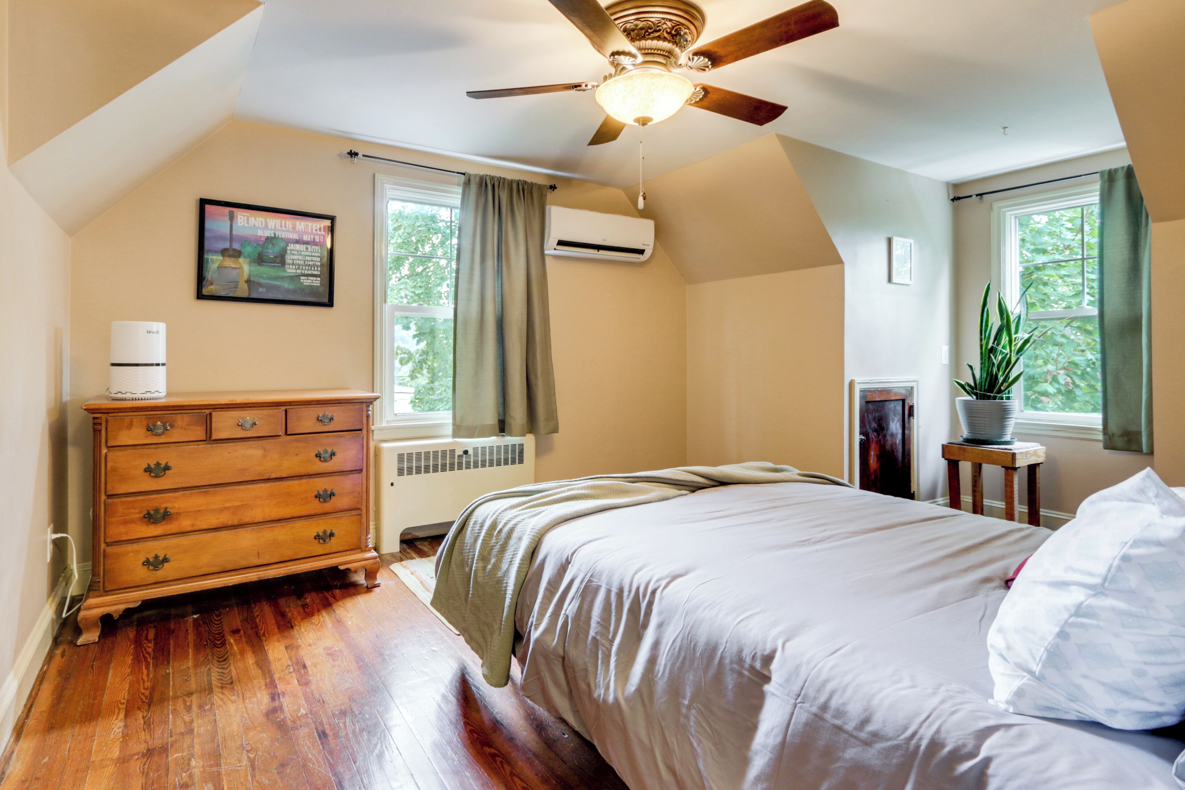 Une chambre à coucher avec une commode en bois, un lit avec une literie blanche, un ventilateur de plafond et une fenêtre avec des rideaux.