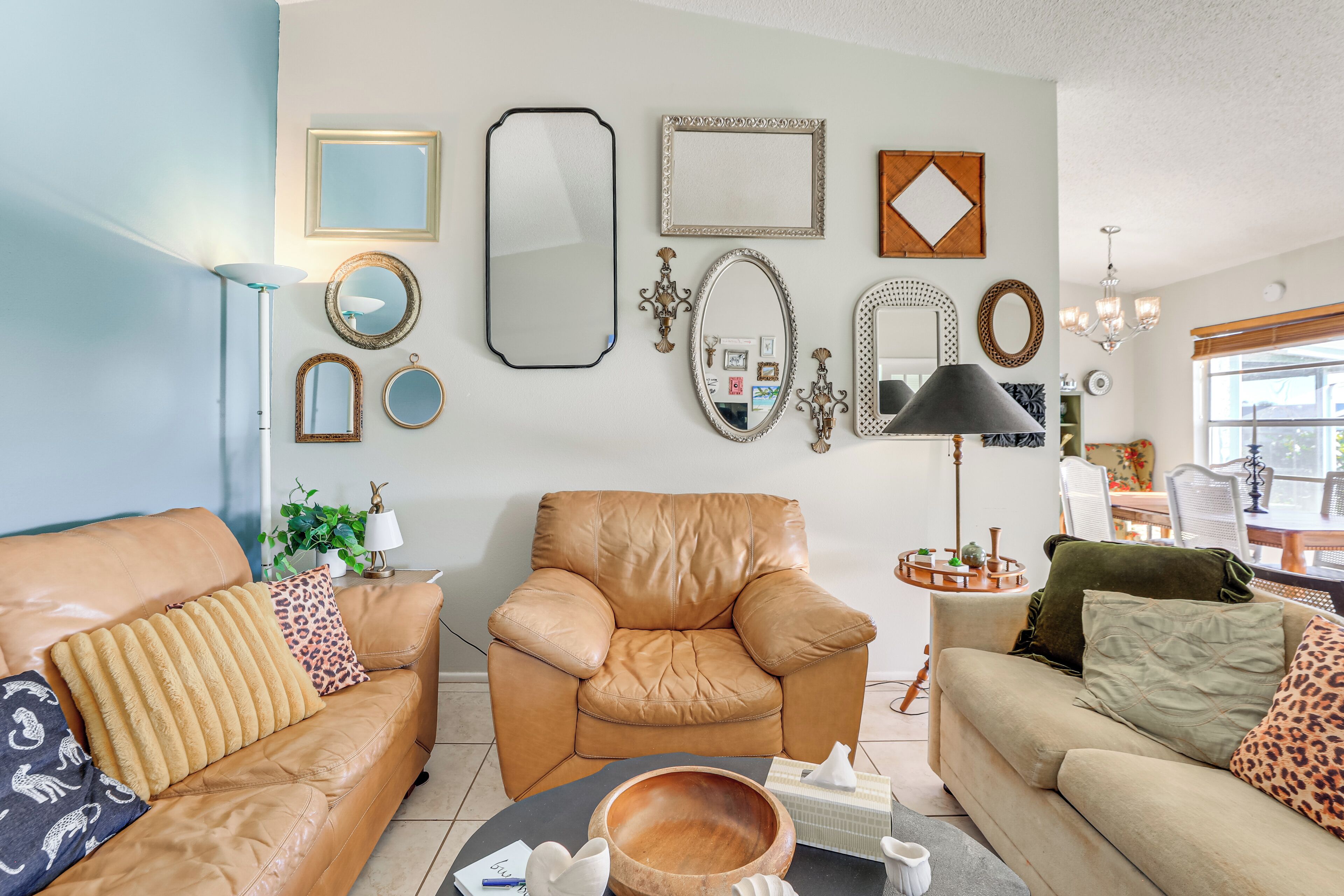 A living room with a brown leather sofa, a matching armchair, and a beige sofa with leopard print pillows. There are various framed mirrors and a chandelier on the wall.
