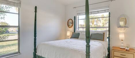 Un lit à baldaquin avec une couvre-lit blanche, une table de chevet avec une lampe et une fenêtre avec des stores.