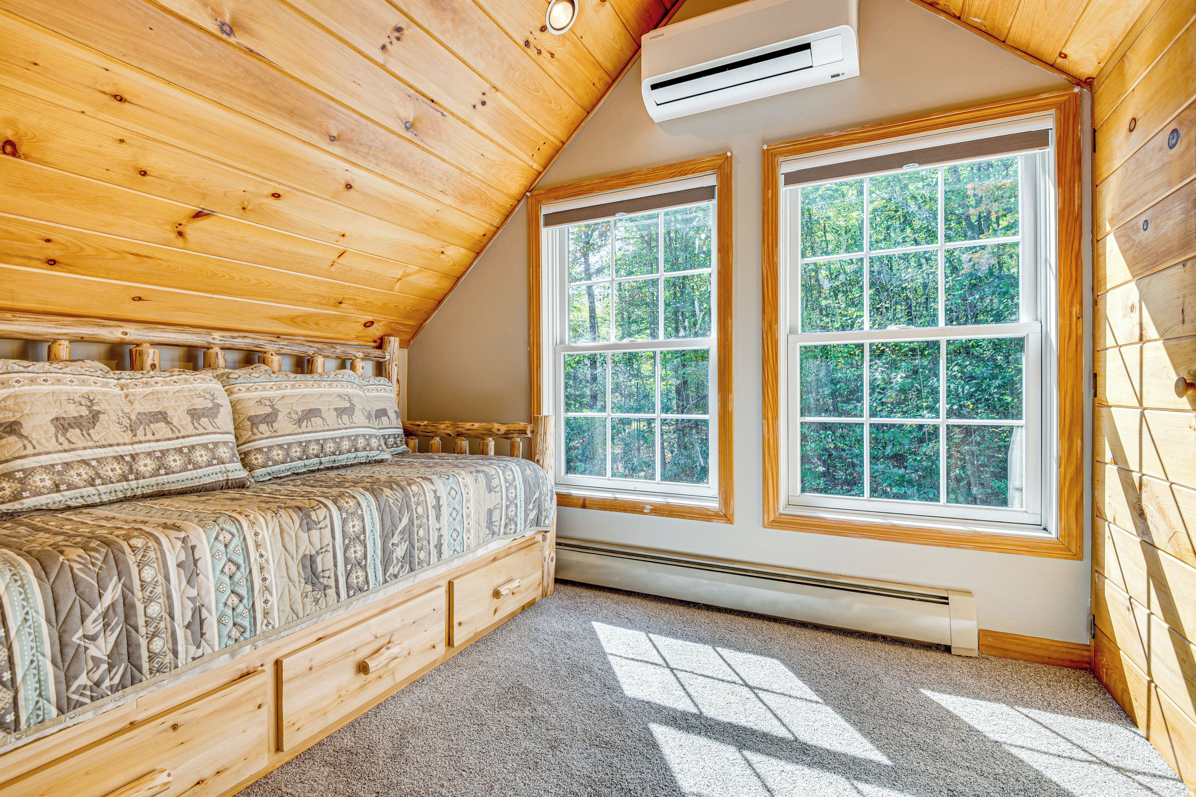 Un dormitorio con techo de madera, dos ventanas grandes, cama con ropa de cama estampada y un banco de almacenamiento integrado.