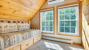 Un dormitorio con techo de madera, dos ventanas grandes, cama con ropa de cama estampada y un banco de almacenamiento integrado.