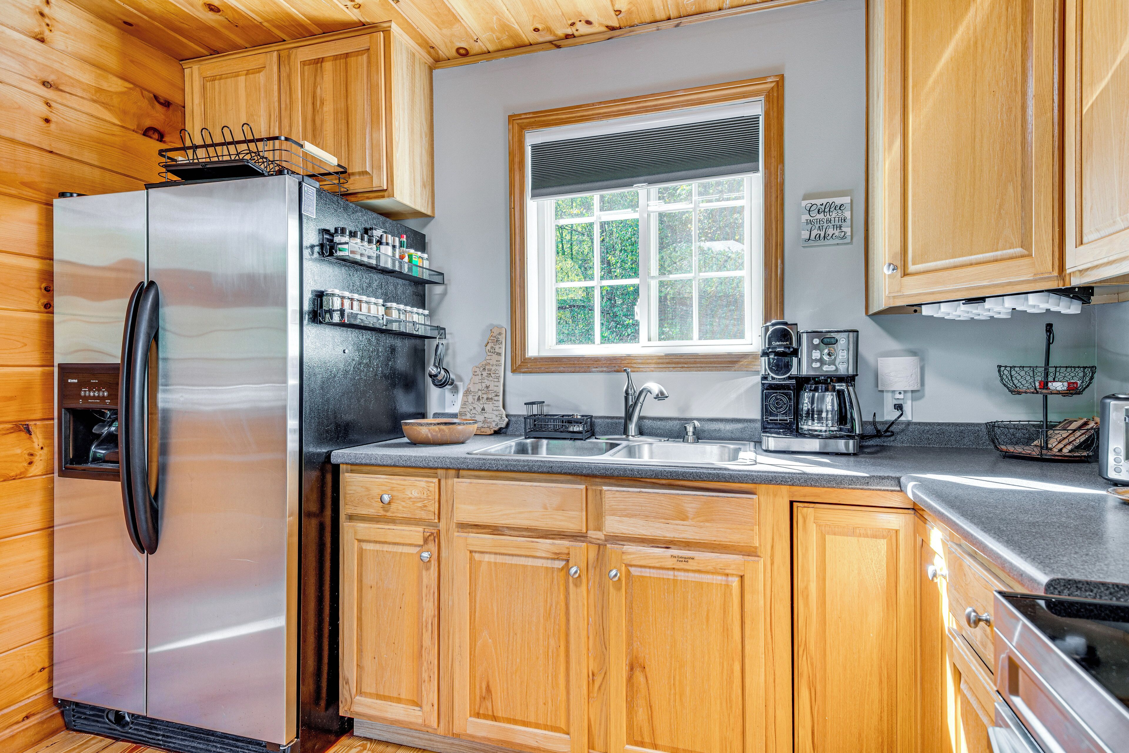 A compact kitchen with wooden cabinets, a stainless steel refrigerator, a coffee maker, and a window with a view of greenery.