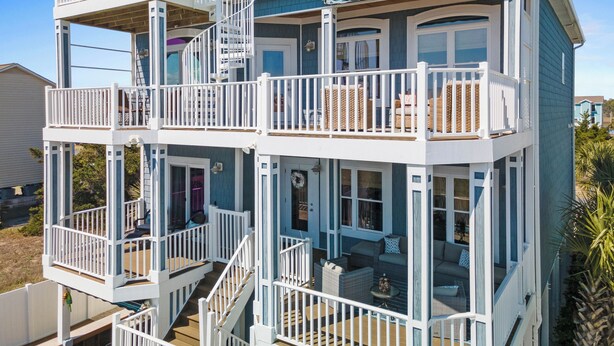 A multi-story house with a balcony on each floor, a staircase connecting the levels, and a porch area with outdoor seating. - Shrimply The Best -Luxury Holden Beach Home with Private Saltwater Pool, Elevator & Tiki Bar (Holden Beach)