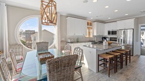 A modern kitchen with white cabinets, a central island with bar stools, and a large window providing a view of the neighborhood. - Shrimply The Best -Luxury Holden Beach Home with Private Saltwater Pool, Elevator & Tiki Bar (Holden Beach)