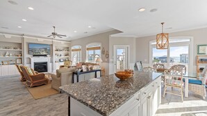 A spacious kitchen with granite countertops, a dining area with wicker furniture, and a living room with a fireplace. - Shrimply The Best -Luxury Holden Beach Home with Private Saltwater Pool, Elevator & Tiki Bar (Holden Beach)