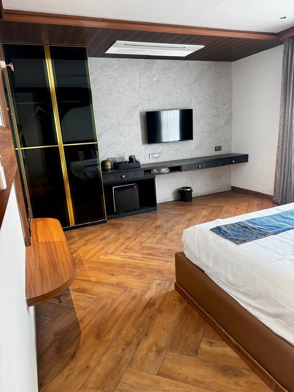 A modern hotel room with a wooden floor, a bed, a TV mounted on the wall, a desk, and a wardrobe with a sliding door.