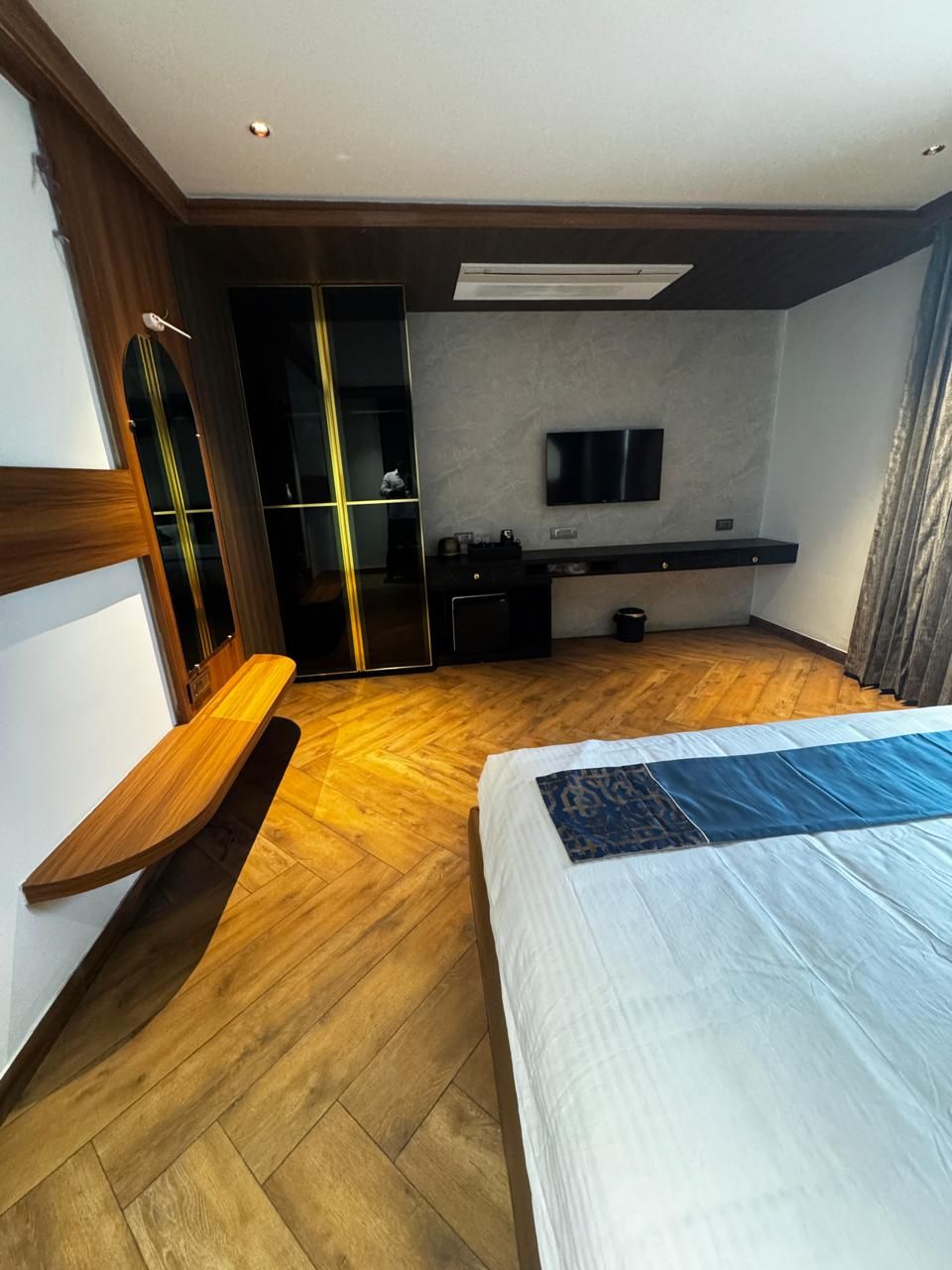 A hotel room with a wooden floor, a bed with a patterned bedspread, a TV mounted on the wall, a desk with a chair, and a sliding door closet.