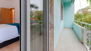 Quarto com cama, porta de vidro deslizante para uma varanda com vista para áreas verdes e edifícios.