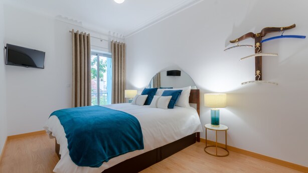 Uma cama bem arrumada com colcha azul e branca, um abajur e uma televisão na parede.