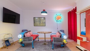 Quarto com duas cadeiras com tema de carros antigos, uma mesinha redonda, um letreiro de neon e uma televisão fixada na parede.