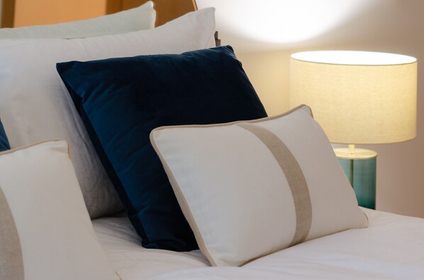 A neatly made bed with white and dark blue pillows, a lamp on a side table, and a wall in the background.