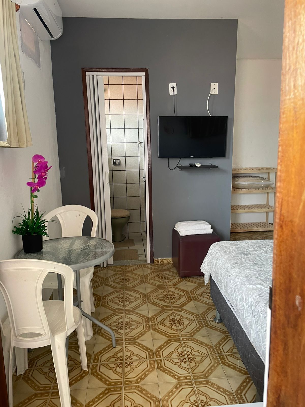 A compact hotel room with a bed, a small table with chairs, a TV mounted on the wall, and a potted plant.