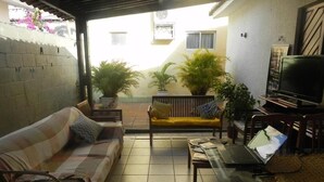 A covered patio with a sofa, a small table, and a laptop. There are potted plants and a TV in the background. - Pousada Zenite (Natal)