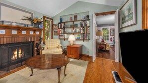 Ein gemütliches Wohnzimmer mit Kamin, einem karierten Sessel, einem Holztisch und einem Bücherregal voller Bücher und Dekoration.