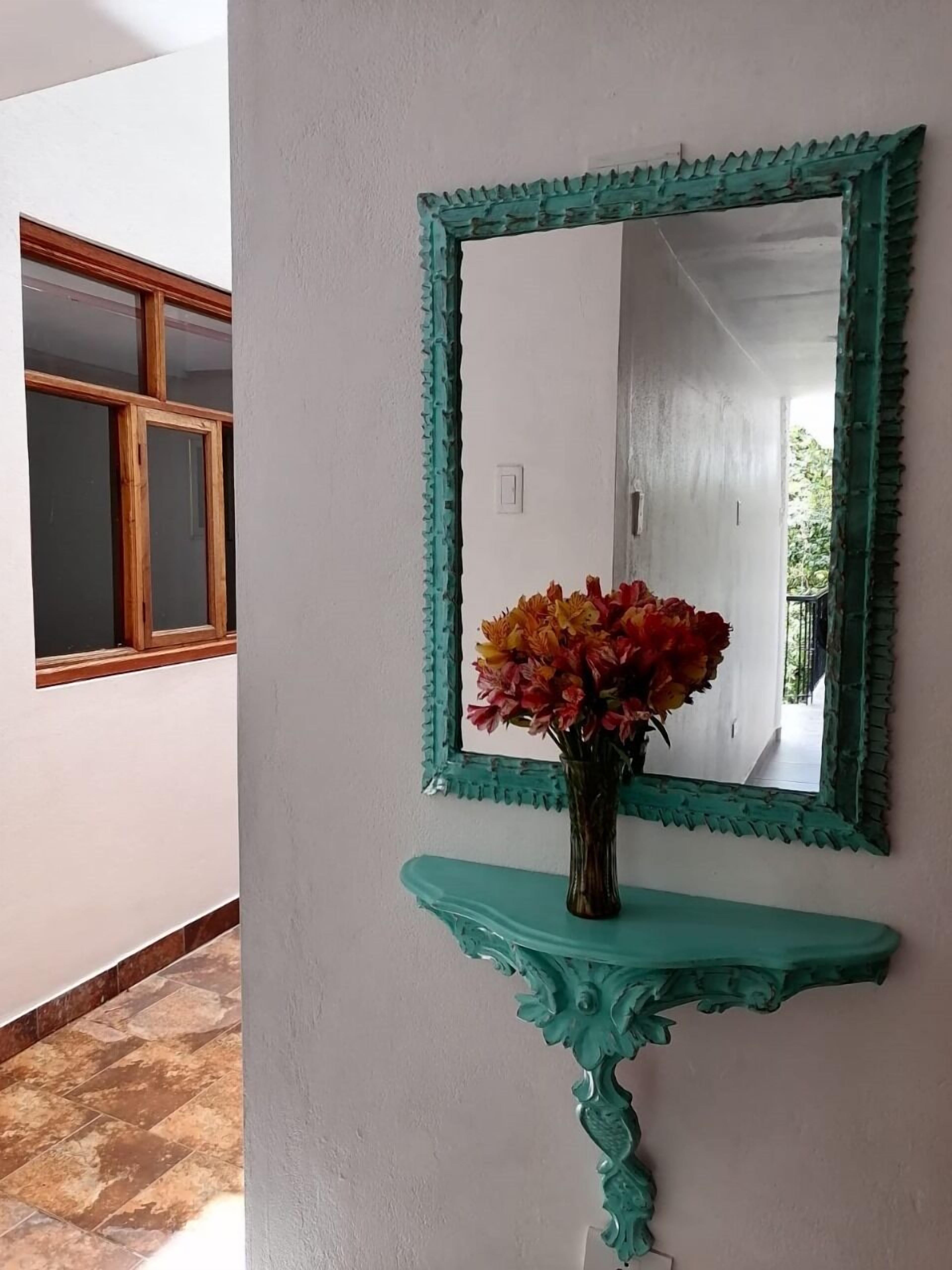 Un couloir avec un miroir placé sur une console de couleur turquoise, une fenêtre en bois et un vase rempli de fleurs.