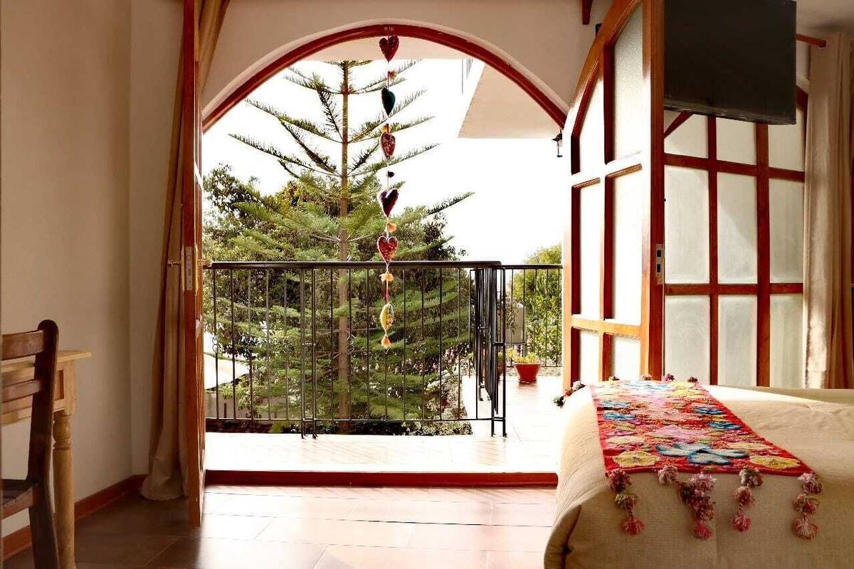 Un balcón con vista a un árbol, una cama con una colcha colorida y una silla de madera.