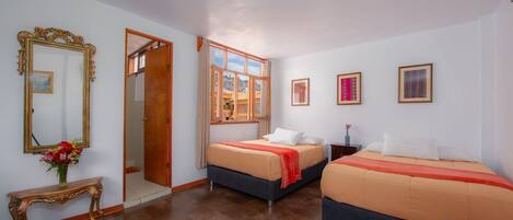 Habitación de hotel con dos camas, piso de madera, espejo, un jarrón de flores y ventana con vistas.