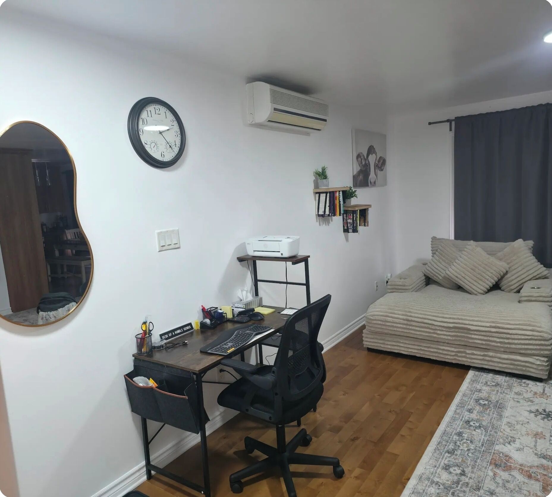 A compact living space with a wooden desk, a black office chair, a round mirror, a clock, an air conditioner, a bookshelf, and a sofa with cushions.