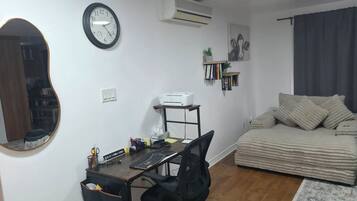 A compact living space with a wooden desk, a black office chair, a round mirror, a clock, an air conditioner, a bookshelf, and a sofa with cushions.