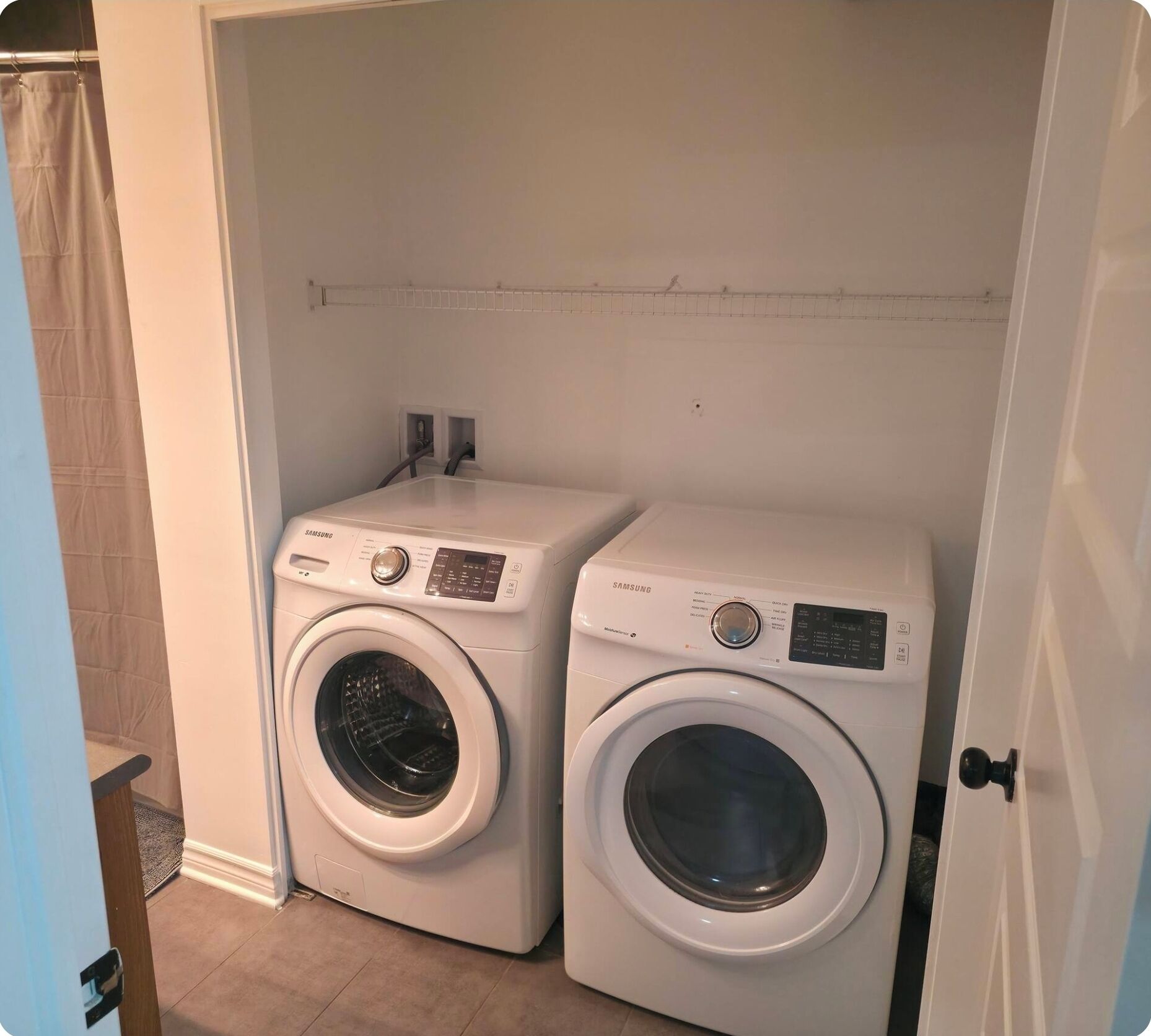 A compact laundry room with two Samsung washing machines, a small shelf, and a door.
