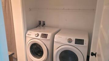 A compact laundry room with two Samsung washing machines, a small shelf, and a door.