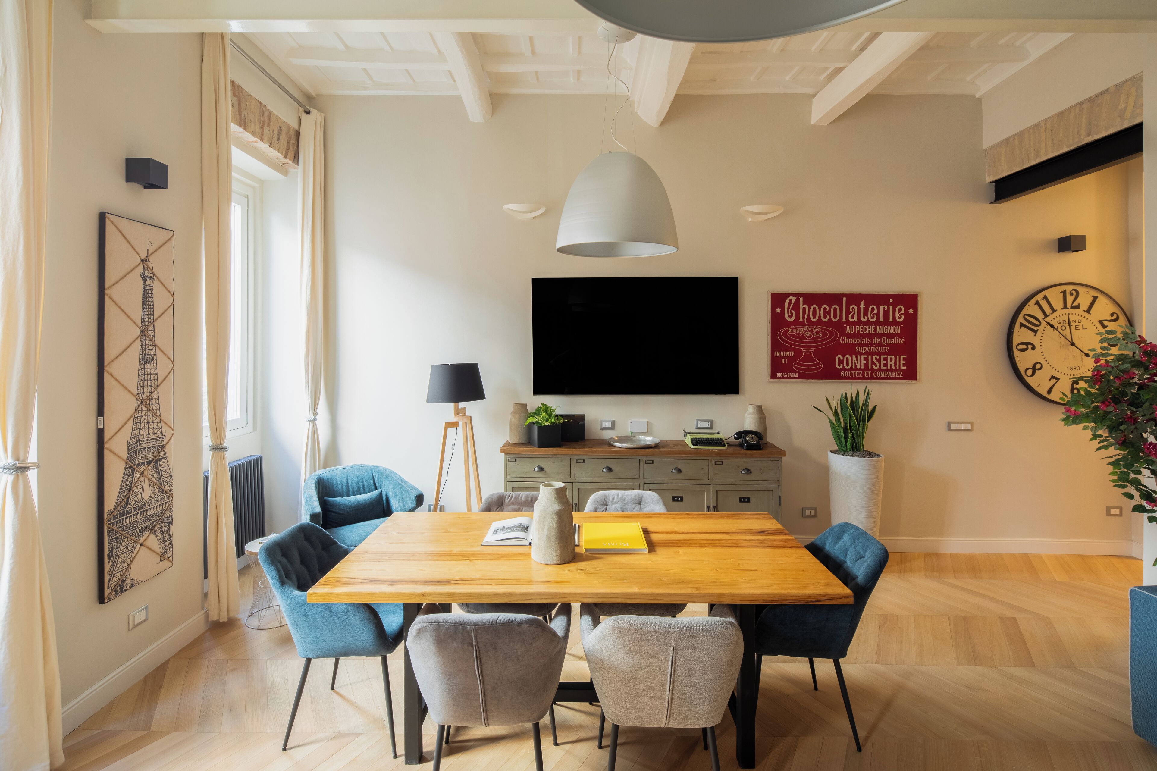 A dining area with a wooden table, blue chairs, a large flat-screen TV, a clock, and a potted plant.