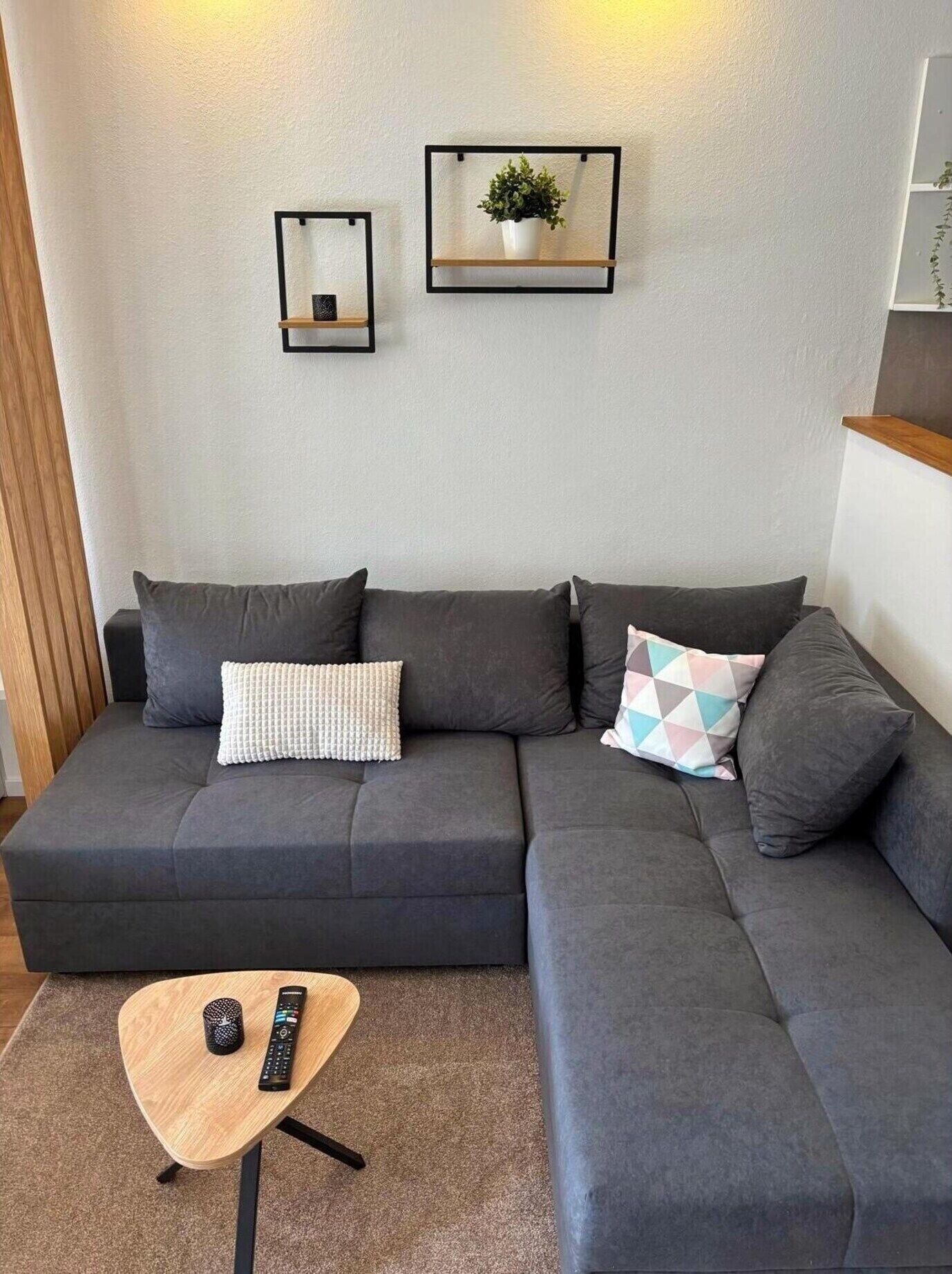 A modern living room with a grey sectional sofa, a small wooden coffee table, a remote control, and two wall-mounted shelves with a plant and a mirror.