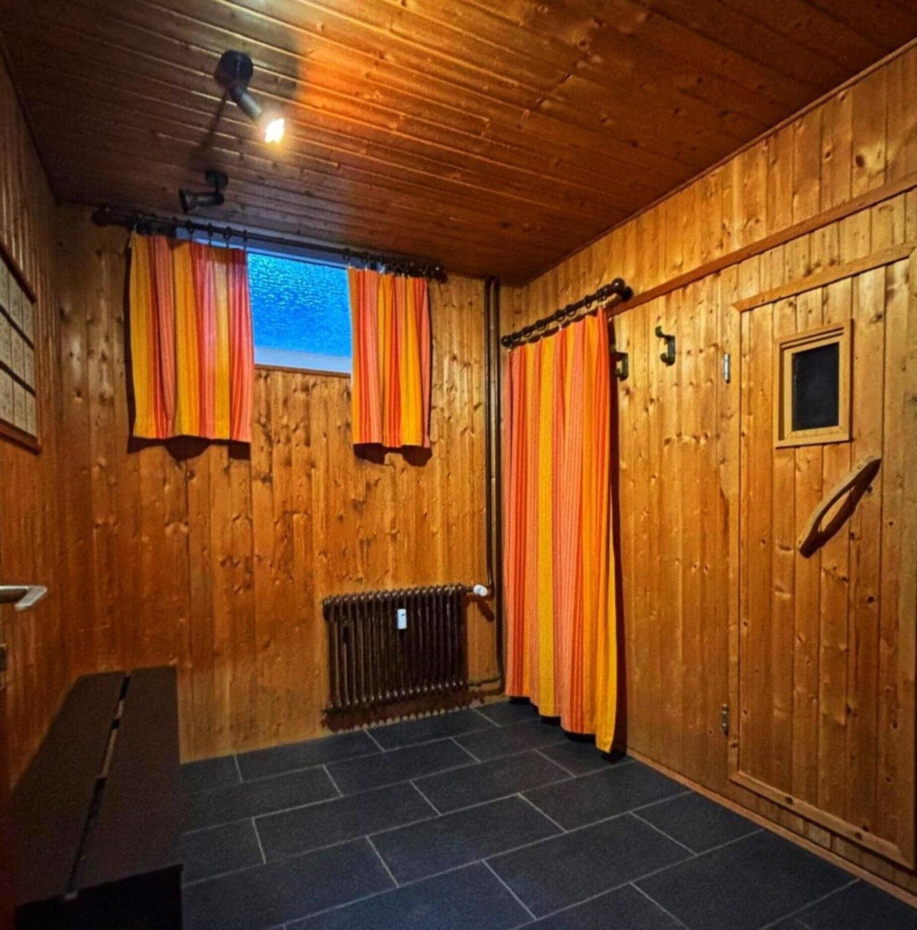 A wooden room with a window, two curtains, a radiator, and a bench.