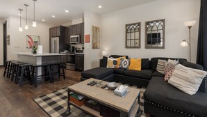 Ein modernes Wohnzimmer mit einer schwarzen Couch, einem Couchtisch und einer Küchenzeile inklusive Kühlschrank und Mikrowelle.