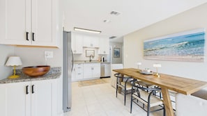 A modern kitchen with white cabinets, a wooden dining table, and a beach painting.