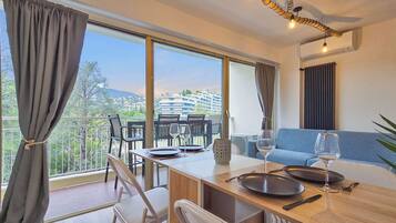 Un comedor con una mesa lista para dos personas, un sofá y un balcón con vistas a edificios y árboles.