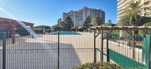 A pool area with a wooden pergola, a fenced pool, and a multi-story building in the background. - LX171 Studio design avec balcon & piscine - Marina (Mandelieu-la-Napoule)