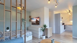 Ein modernes Wohnzimmer mit einer Couch, einem Couchtisch und einem an der Wand montierten Fernseher. Eine Treppe führt zu einer oberen Ebene, und im Hintergrund ist eine Küchenzeile zu sehen.