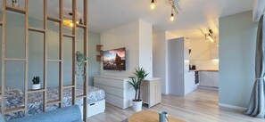 A modern living room with a sofa, a coffee table, and a TV mounted on the wall. There is a staircase leading to an upper level, and a kitchen area is visible in the background. - LX171 Studio design avec balcon & piscine - Marina (Mandelieu-la-Napoule)
