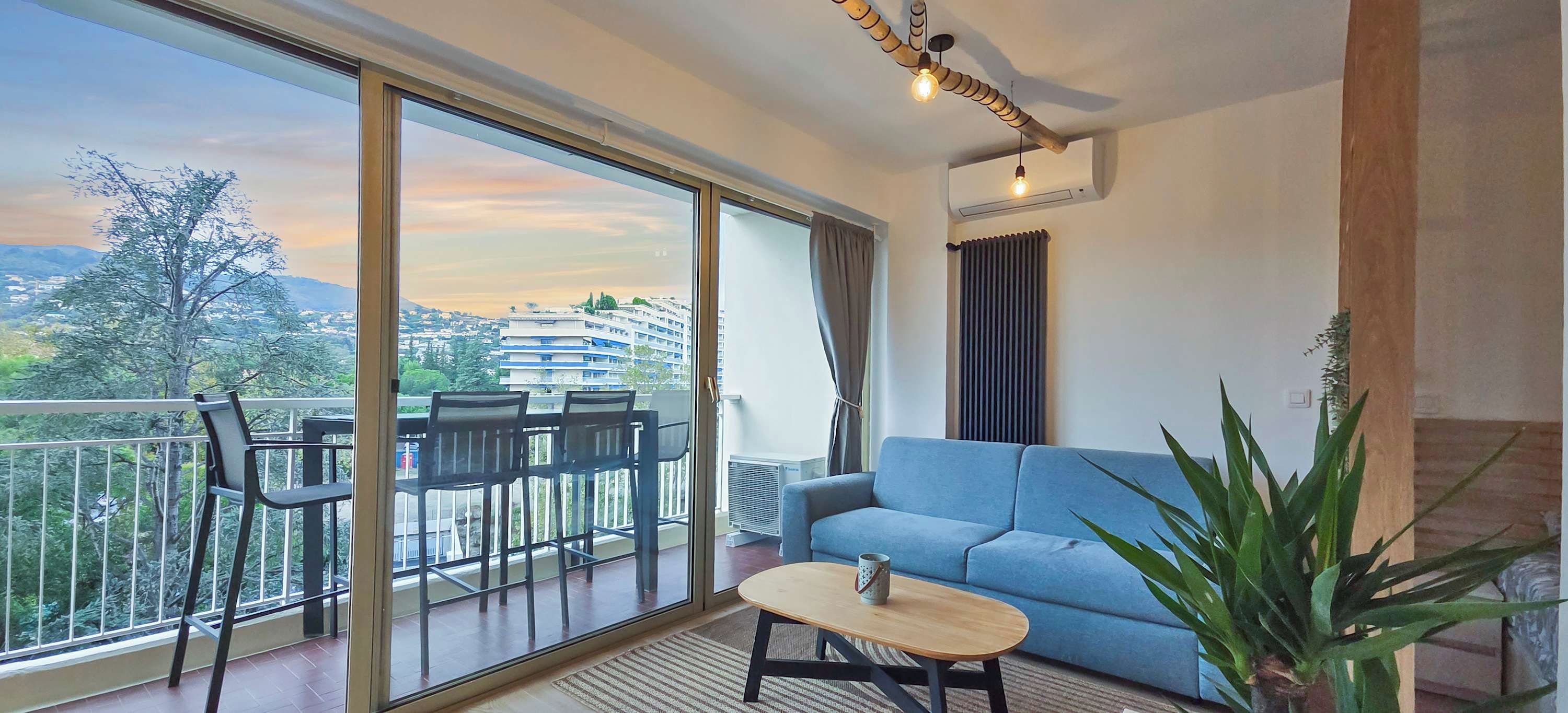 A modern living room with a blue sofa, a wooden coffee table, and a balcony with chairs and a view of the city and mountains.