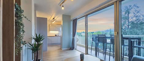 A modern apartment with a kitchen, a balcony with chairs, and large windows offering a view of the city.