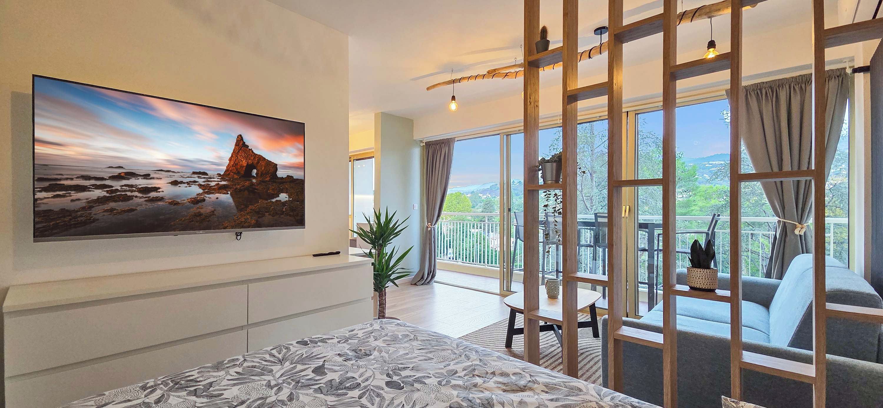 Una moderna camera d'hotel con una grande TV a schermo piatto montata a parete, un letto con copripiumino decorato, un balcone che offre vista sul paesaggio e un pannello in legno con una lampada a sospensione.